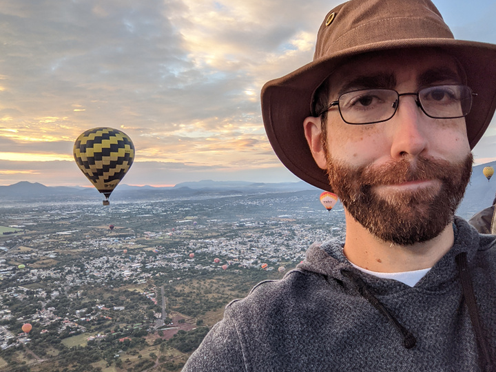 A selfie of me, con sombrero, looking unamused from 1000 feet off the ground, with a sunrise, mountains, and other hot air balloons behind me.